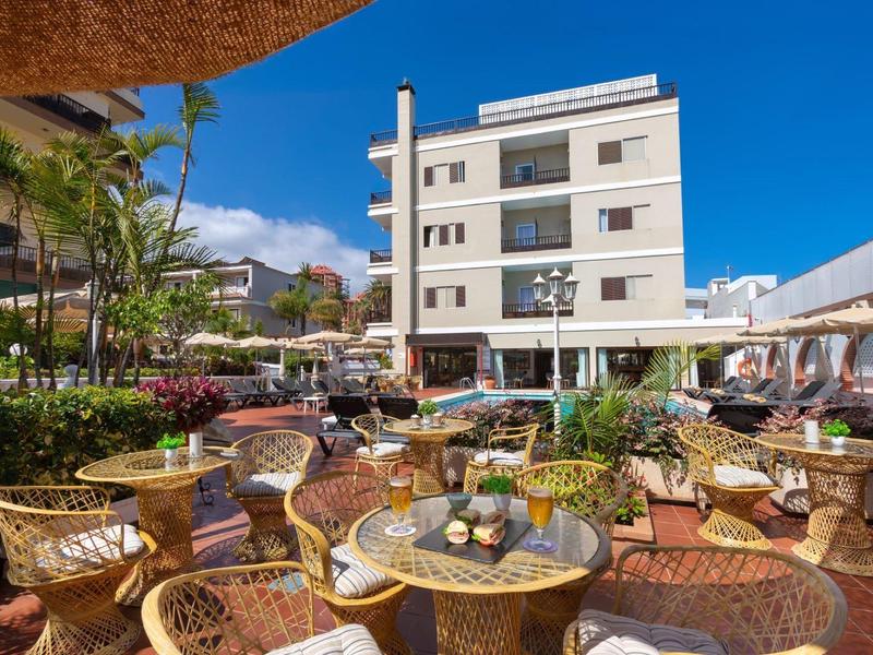 Buitenloungezitplaatsen met tafels en planten voor een meerlaagse hotel onder een blauwe lucht.