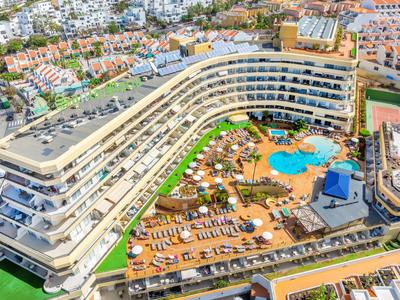 Grande hotel curvo con piscina, terrazza solarium e lettini in una località turistica soleggiata.