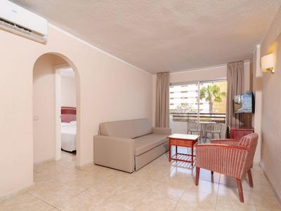 Soggiorno luminoso con divano, poltrone, TV e balcone con vista su tetti e palme.