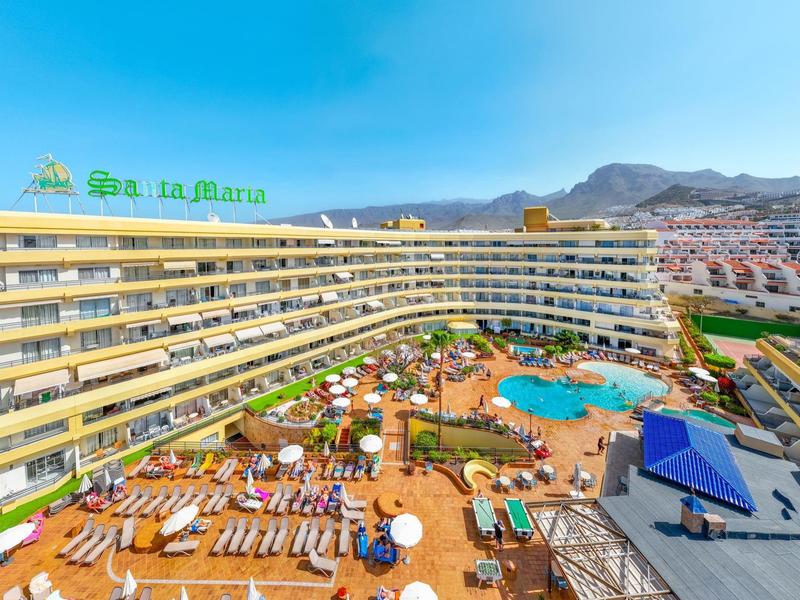 Vista di un grande hotel su più piani con piscina e molte sdraio sotto un cielo azzurro.