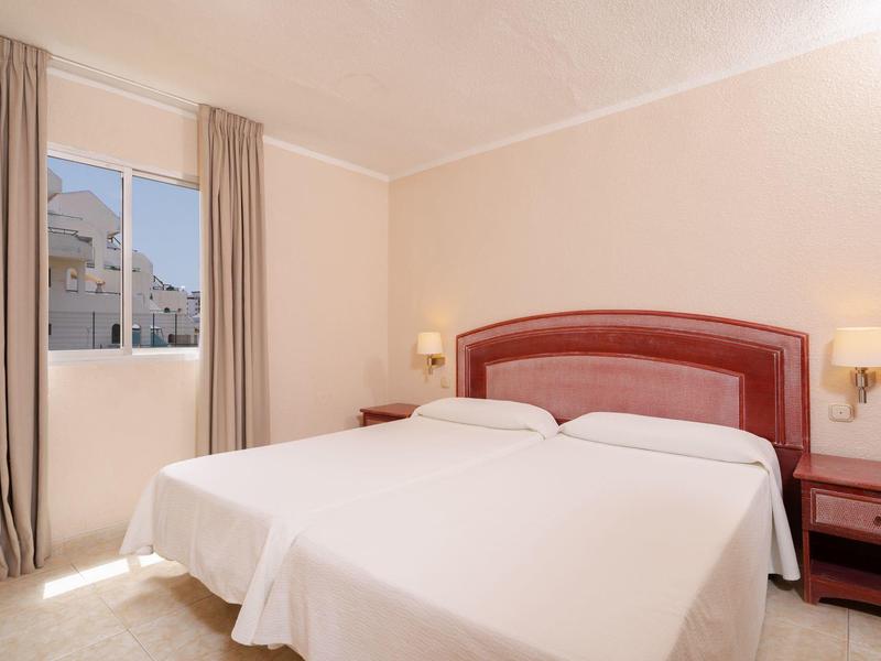 Camera d'hotel luminosa con due letti singoli, comodini rossi e finestre con tende.