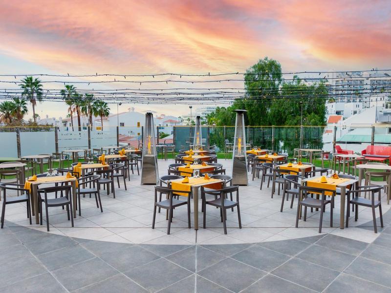 Area ristorante aperta con tavoli e sedie sotto un cielo colorato al tramonto.