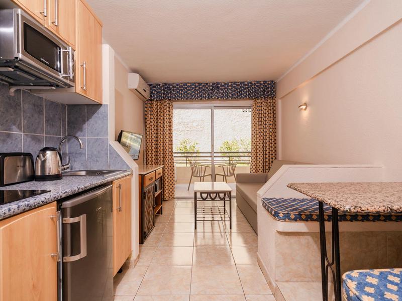 Cucina compatta con mobili in legno, tavolo da pranzo e divano accanto a una finestra con vista su un balcone.