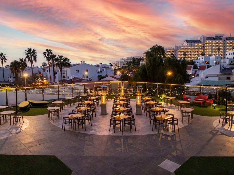 Vista esterna serale di un ristorante illuminato con sedie e tavoli sotto un cielo colorato.