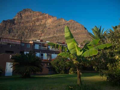 Haus mit braunen Wänden vor großem Felsen, grüner Wiese und Palmen unter klarem blauem Himmel.