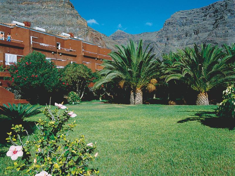 Grüner Garten mit Palmen, rosa Blumen und mehrstöckigem Gebäude vor felsiger Berglandschaft.