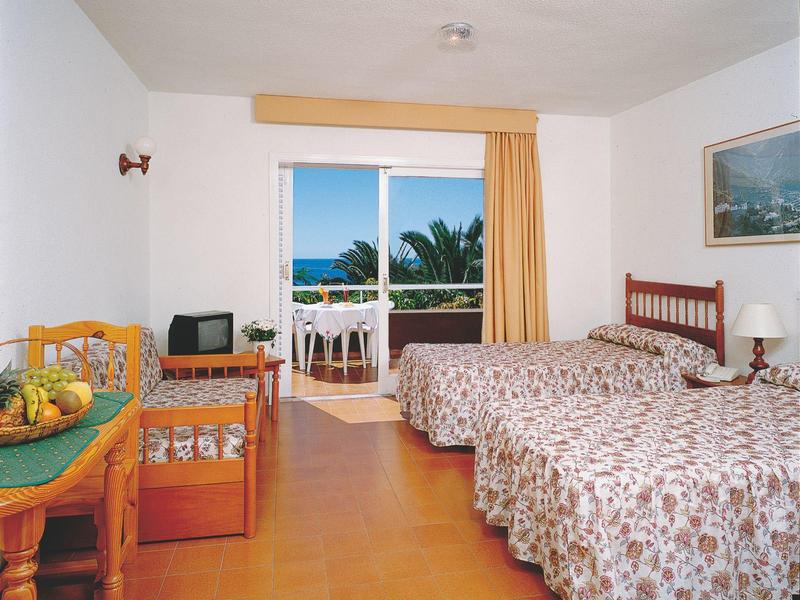 Helles Zimmer mit zwei Einzelbetten, großem Fenster, Holzmöbeln und Balkon mit Meerblick.