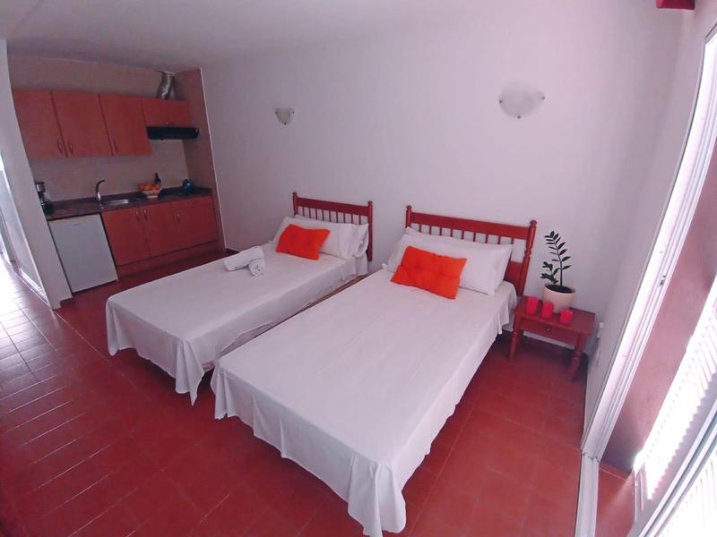 Einfaches Zimmer mit zwei weißen Einzelbetten, roten Kopfteil und Kissen, kleine Küchenzeile links.