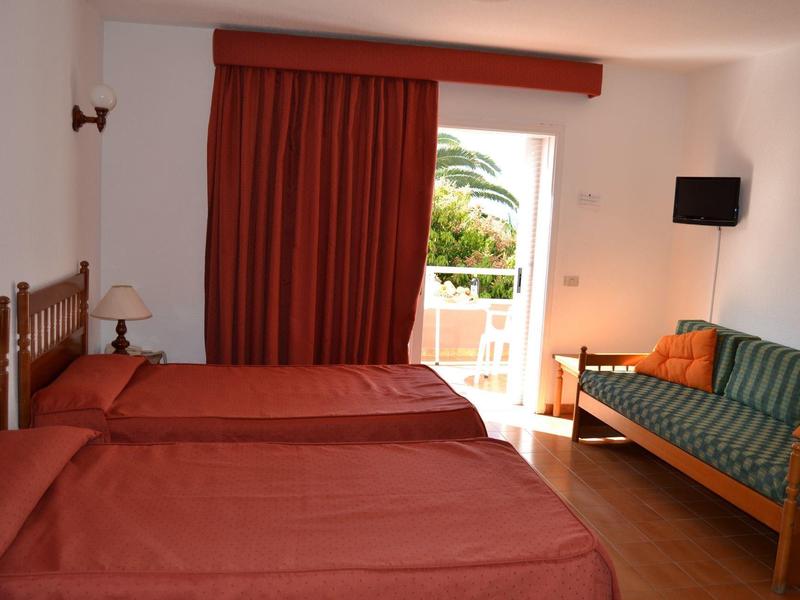 Zimmer mit zwei Einzelbetten, roter Bettwäsche, Sofa mit grün-orangem Muster und Balkonblick.