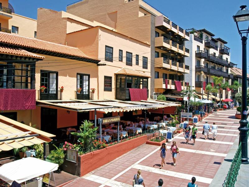 Belebte Fußgängerzone mit Cafés, mehrstöckigen Gebäuden und Menschen bei Sonnenschein.