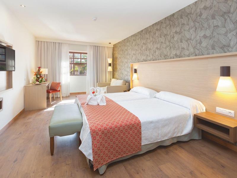 Hell eingerichtetes Hotelzimmer mit Doppelbett, Teppich, Fernseher an der Wand und einem Fenster.