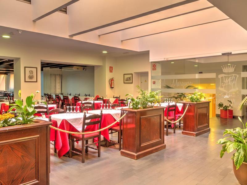 Heller Speiseraum mit weißen Tischen, roten Tischdecken, Holzmöbeln und grünen Pflanzen.