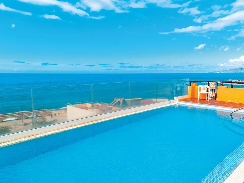 Ein blauer Pool vor blauem Himmel und Meer mit einigen weiß-roten Häusern im Hintergrund.