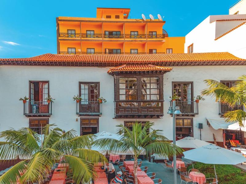 Mehrstöckiges weißes Gebäude mit Holzfenstern und roten Dachziegeln, davor Palmen und Stühle.