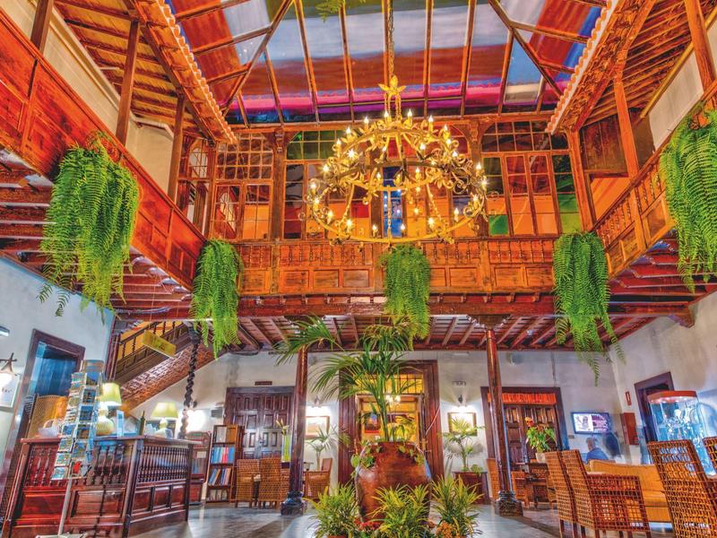 Großer, heller Gastraum mit Holzdecke, vielen Pflanzen und goldenem Kronleuchter in warmen Farben.