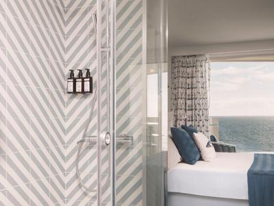Camera d'hotel con doccia in vetro, letto moderno, accenti blu e vista sul mare.