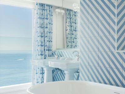 Bagno luminoso con vasca autoportante, piastrelle a righe blu e bianche e vista sul mare.