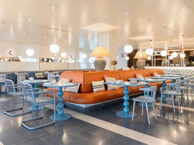 Ristorante d'hotel moderno con divano arancione, tavoli e sedie blu sotto lampade bianche.