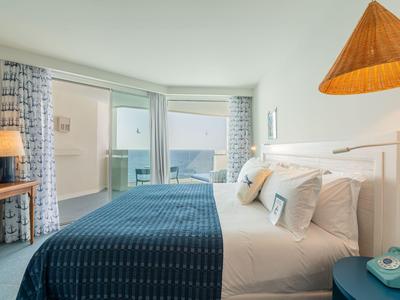 Camera d'hotel luminosa con letto grande, copriletto blu e vista mare dalla porta del balcone.