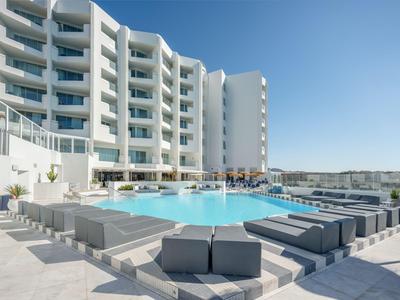 Hotel moderno con facciata bianca e area piscina con mobili lounge grigi sotto un cielo limpido.