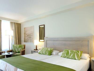 Jasny pokój hotelowy z podwójnym łóżkiem, zielonymi poduszkami, dwoma fotelami i dużym oknem.