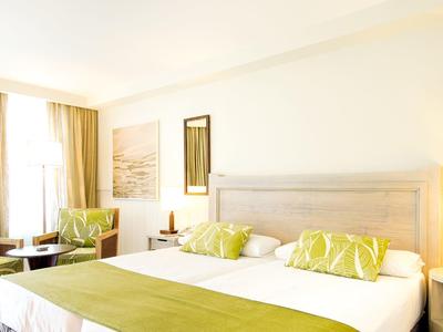 Jasny pokój hotelowy z podwójnym łóżkiem, zielonymi akcentami, dwoma fotelami i dużym oknem z zasłonami.