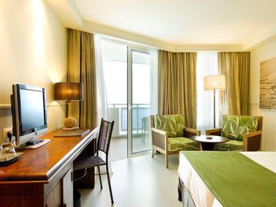 Jasny pokój hotelowy z łóżkiem, biurkiem, telewizorem, fotelem i balkonem z przezroczystymi zasłonami.