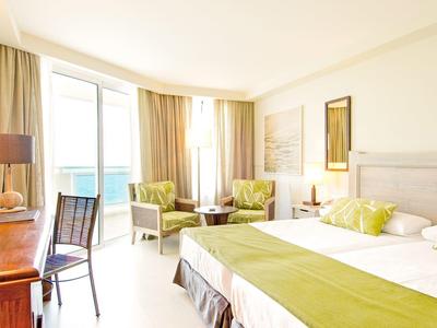 Jasny pokój hotelowy z podwójnym łóżkiem, biurkiem, fotelami i widokiem na morze przez duże okna.