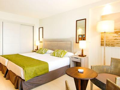 Jasny pokój hotelowy z dwoma łóżkami, zielonymi akcentami, fotelami i małym stolikiem.