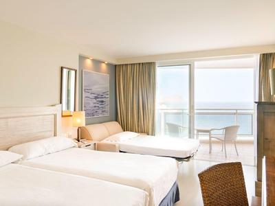 Jasny pokój hotelowy z dwoma łóżkami, balkonem z widokiem na morze i neutralnym wystrojem.