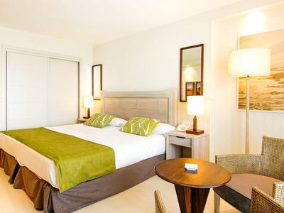 Jasny pokój hotelowy z dużym łóżkiem, zielonymi akcentami, fotelami i ciepłym oświetleniem.