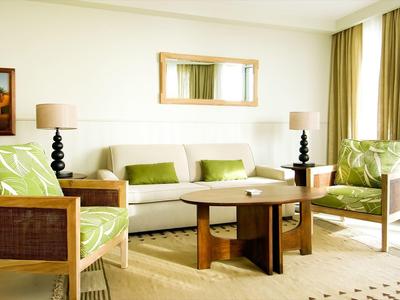 Jasny salon hotelowy z sofą, fotelami, drewnianym stolikiem i dużymi oknami z zasłonami.