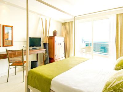 Jasny pokój hotelowy z dużym łóżkiem, zielonymi akcentami, biurkiem, telewizorem i balkonem z przeszklonymi drzwiami.