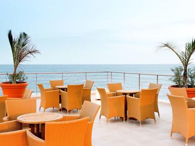 Zewnętrzny teren wypoczynkowy z pomarańczowymi krzesłami i stołami z widokiem na morze i palmy.