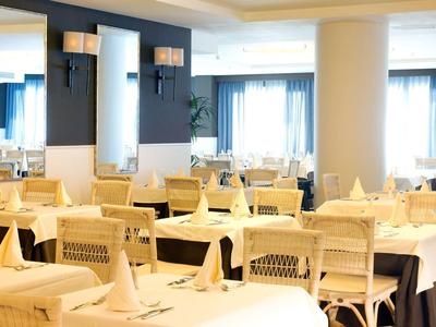 Jasna sala jadalna hotelu z białymi obrusami, złożonymi serwetkami i nowoczesnymi drewnianymi krzesłami.