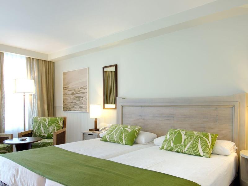 Jasny pokój hotelowy z podwójnym łóżkiem, zielonymi poduszkami, dwoma fotelami i dużym oknem.