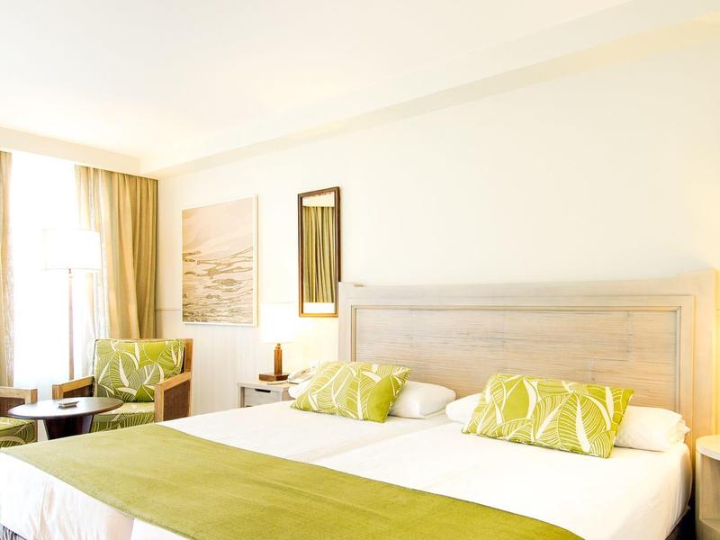 Jasny pokój hotelowy z podwójnym łóżkiem, zielonymi akcentami, dwoma fotelami i dużym oknem z zasłonami.
