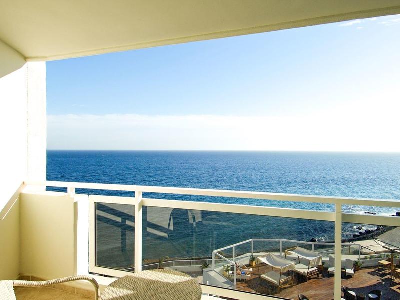 Balkon z szklaną barierką z widokiem na niebieskie morze i czyste niebo, z miejscami do siedzenia na zewnątrz.