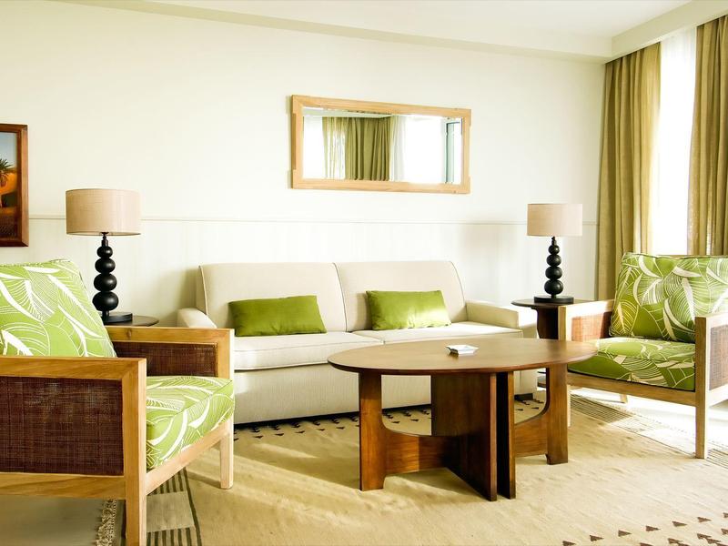 Jasny salon hotelowy z sofą, fotelami, drewnianym stolikiem i dużymi oknami z zasłonami.