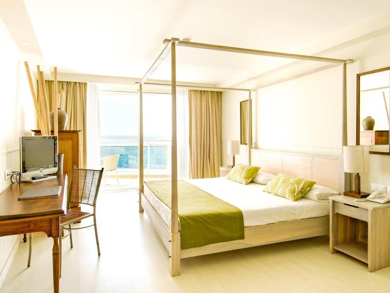 Jasny pokój hotelowy z łóżkiem z baldachimem, drewnianym biurkiem i balkonem z widokiem na ocean.