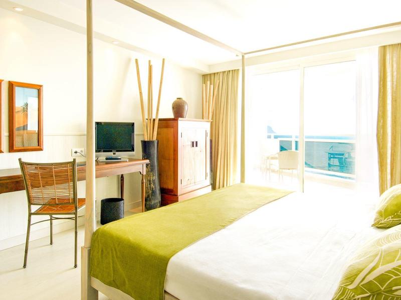 Jasny pokój hotelowy z dużym łóżkiem, zielonymi akcentami, biurkiem, telewizorem i balkonem z przeszklonymi drzwiami.