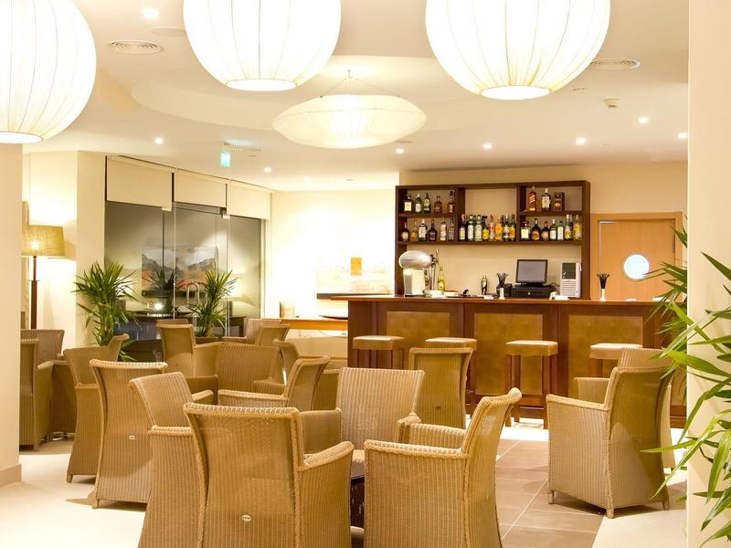 Przytulny salon hotelowy z wiklinowymi krzesłami, okrągłymi stolikami, roślinami i barem przy ciepłym oświetleniu.