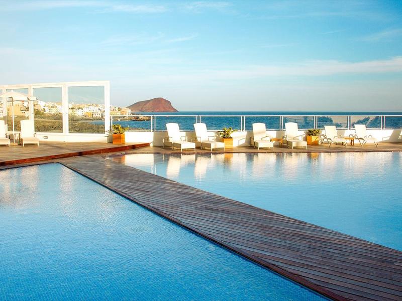 Duży basen na zewnątrz z drewnianym pomostem i białymi leżakami nad morzem pod czystym niebem.