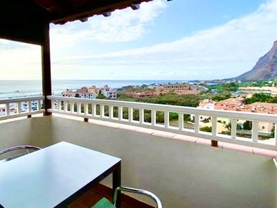 Balkon mit Tisch und Stühlen, Blick auf Meer, Ort und Berge bei klarem Himmel.
