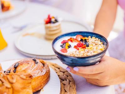 Osoba drží misku s jogurtem, granolou a bobulemi vedle pečiva na stole.
