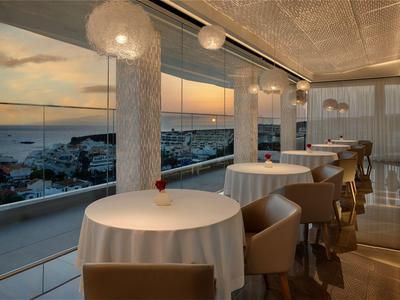 Elegantní restaurace s kulatými stoly a výhledem na město při západu slunce.