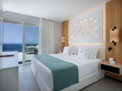 Moderní ložnice s manželskou postelí, výhledem na moře a balkonem v hotelovém pokoji.