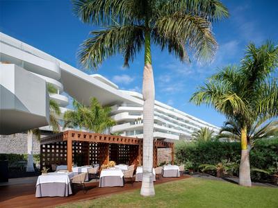 Venkovní prostor moderního hotelu s bílými stoly pod palmami a modrou oblohou.