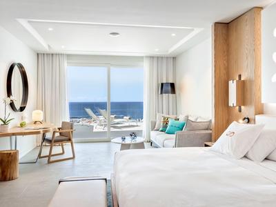 Moderní hotelový pokoj s výhledem na moře, bílými zdmi, dřevěnými prvky a stylovým zařízením.