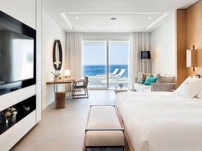 Modern hotel room with large bed, TV, desk, and balcony access overlooking the sea.